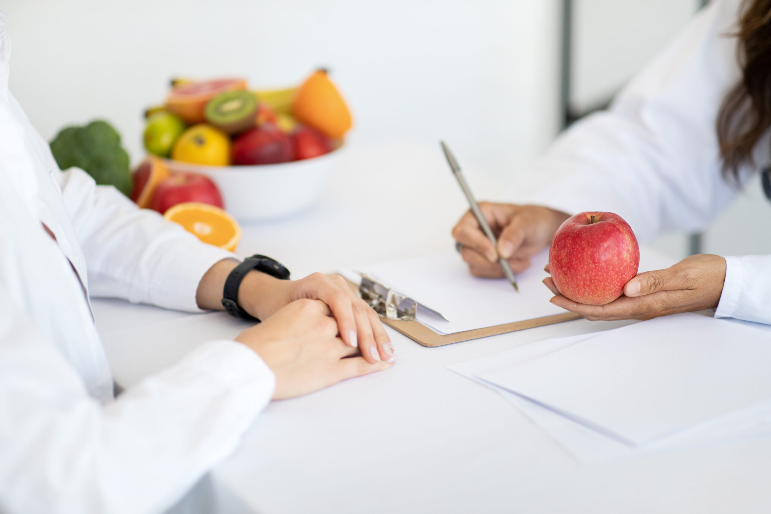 European middle aged lady doctor nutritionist in white coat write diet program to woman patient, recommends apple